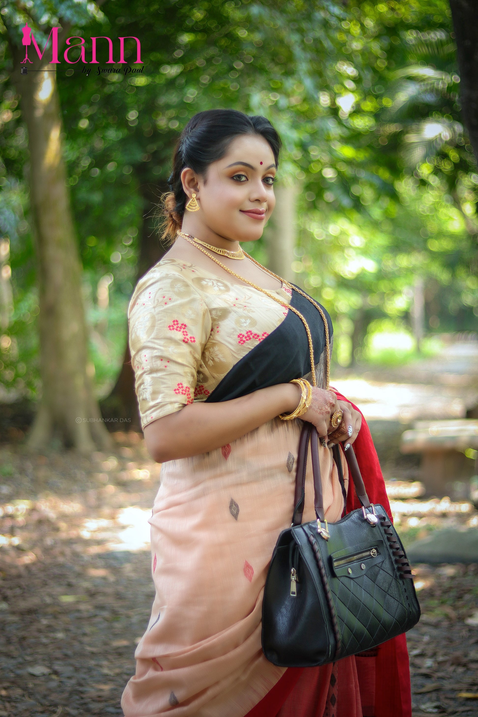 Side view of model wearing a beige semi-silk blouse with subtle floral embroidery in red and gold, paired with a peach and black bordered saree, accessorized with gold jewelry and a black leather handbag, posing in a sunlit forest path