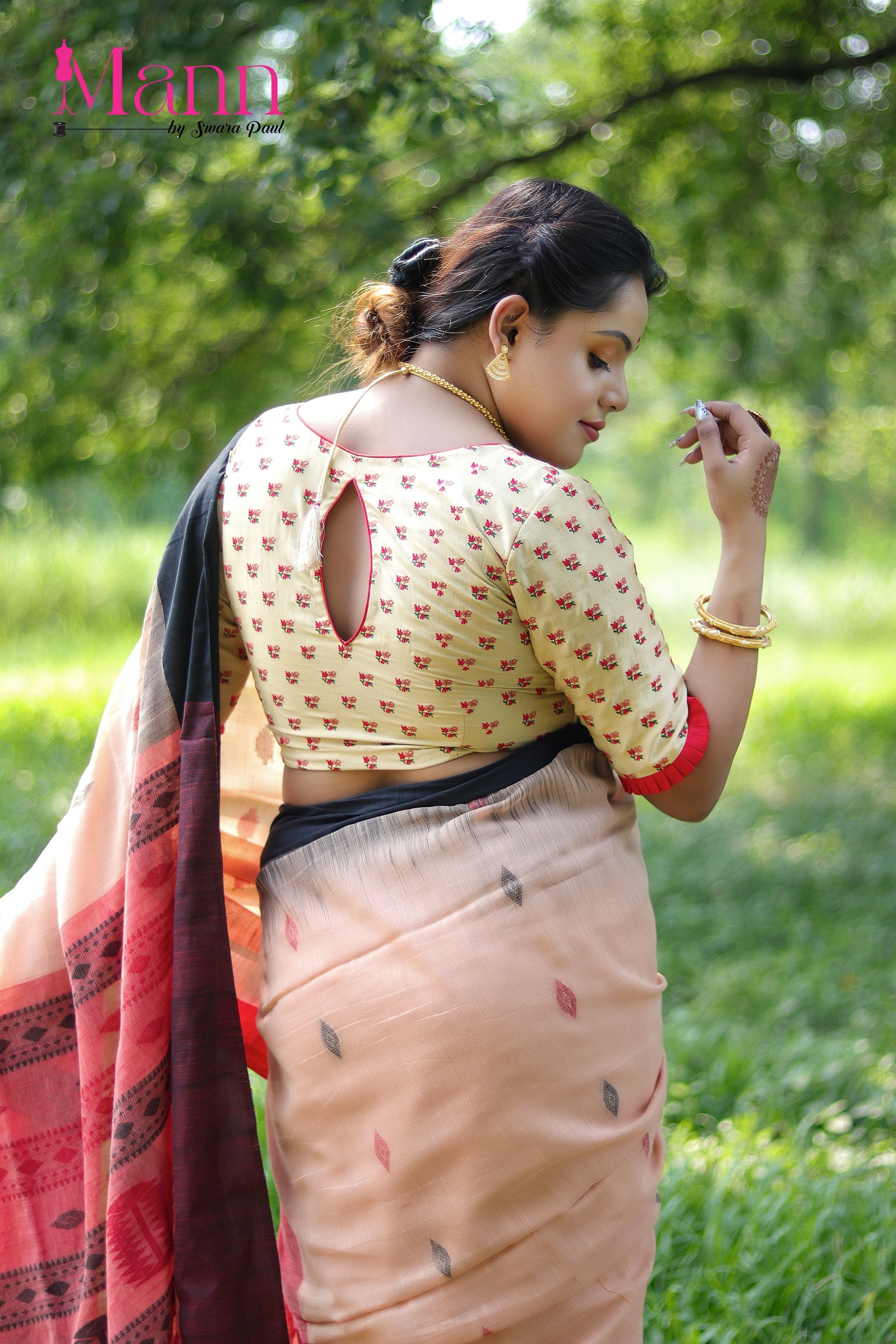 Rear view of beige semi-silk blouse featuring fine red floral threadwork and a teardrop back cutout with tassel tie, styled with a peach saree and bold pleated sleeve edging, model striking a graceful pose with hand near face in an outdoor garden setting