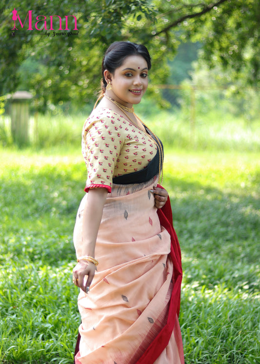 Side view of beige semi-silk blouse with red floral thread embroidery and wide V neckline, paired with a peach and red bordered saree, model posing outdoors with traditional jewellery