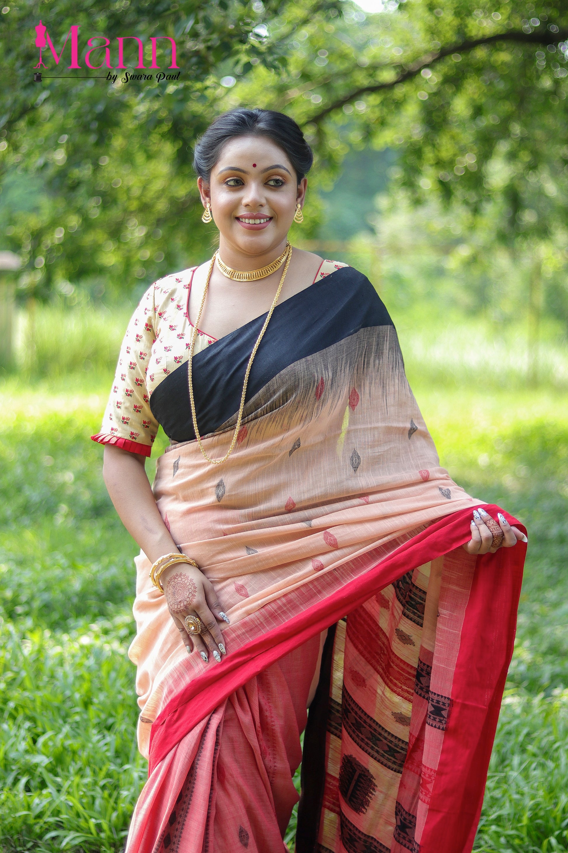 Front view of beige semi-silk blouse with red floral embroidery and wide V neckline, paired with a peach and black bordered saree, model posing outdoors with traditional gold jewellery and mehendi
