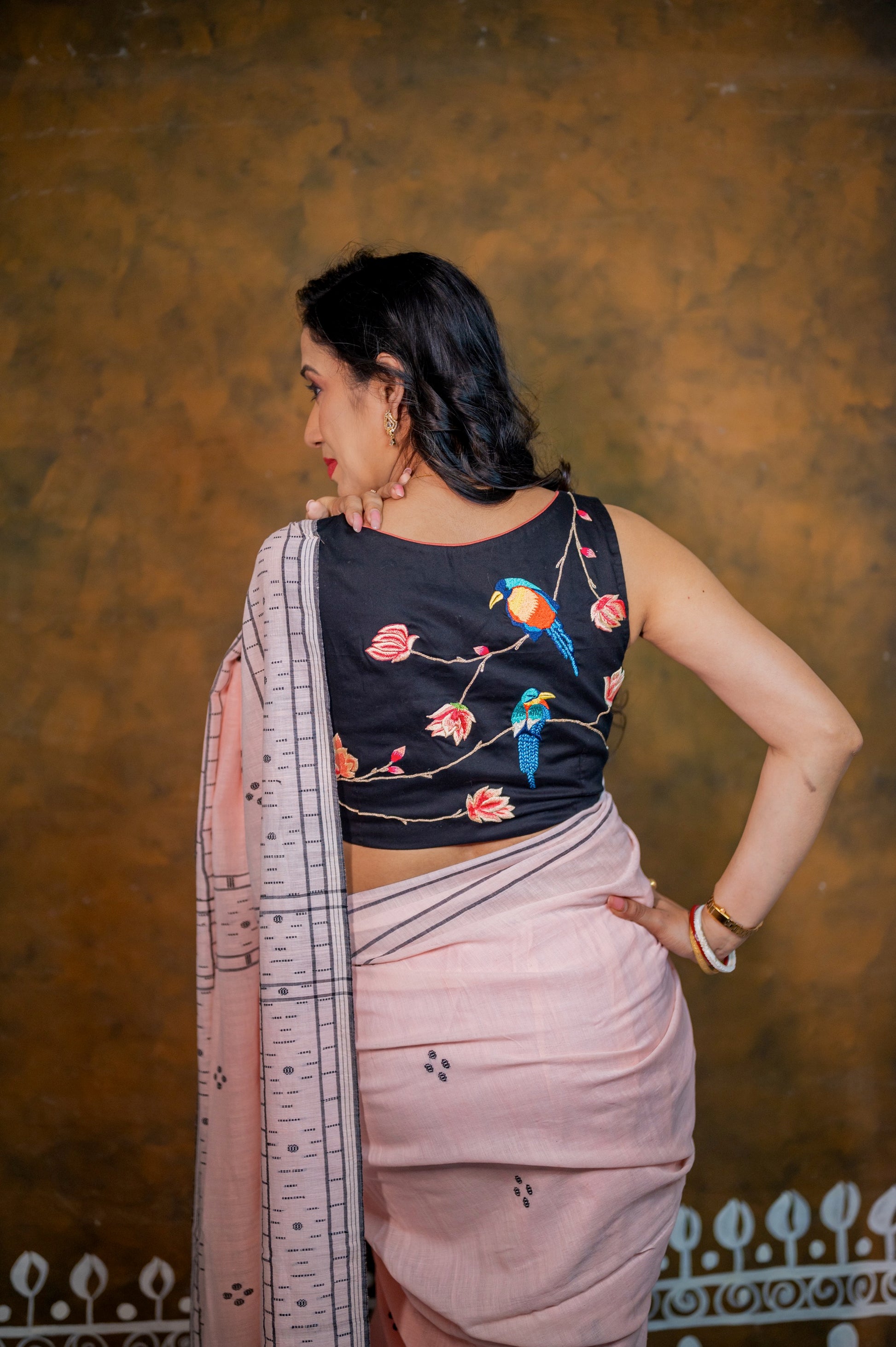 Back view of a sleeveless black blouse with vibrant bird and floral embroidery, paired with a soft pink saree.