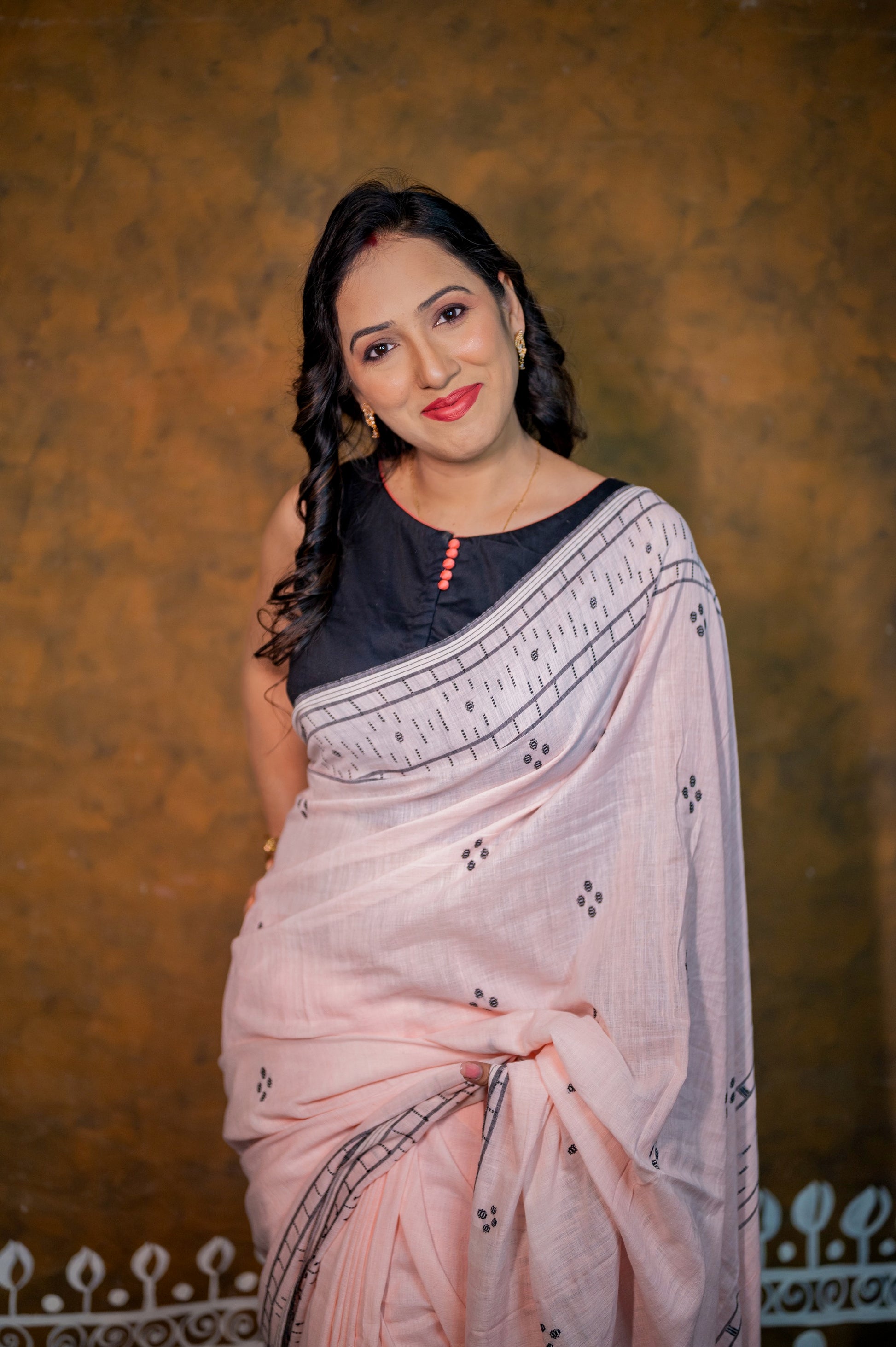 Front view of a black sleeveless blouse with coral pink buttons, styled with a light pink handloom saree.