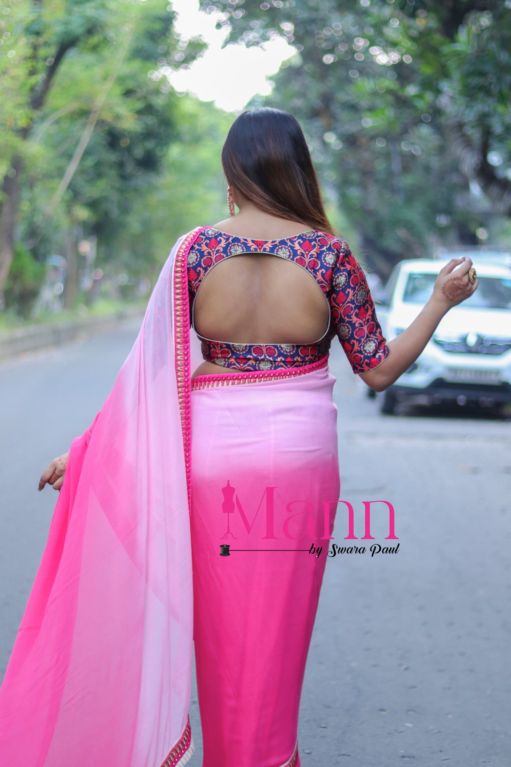 Back view of Banarasi blouse with rich multicolor motifs and curved designer cut-out, styled with a pink ombré saree, model walking down a tree-lined street