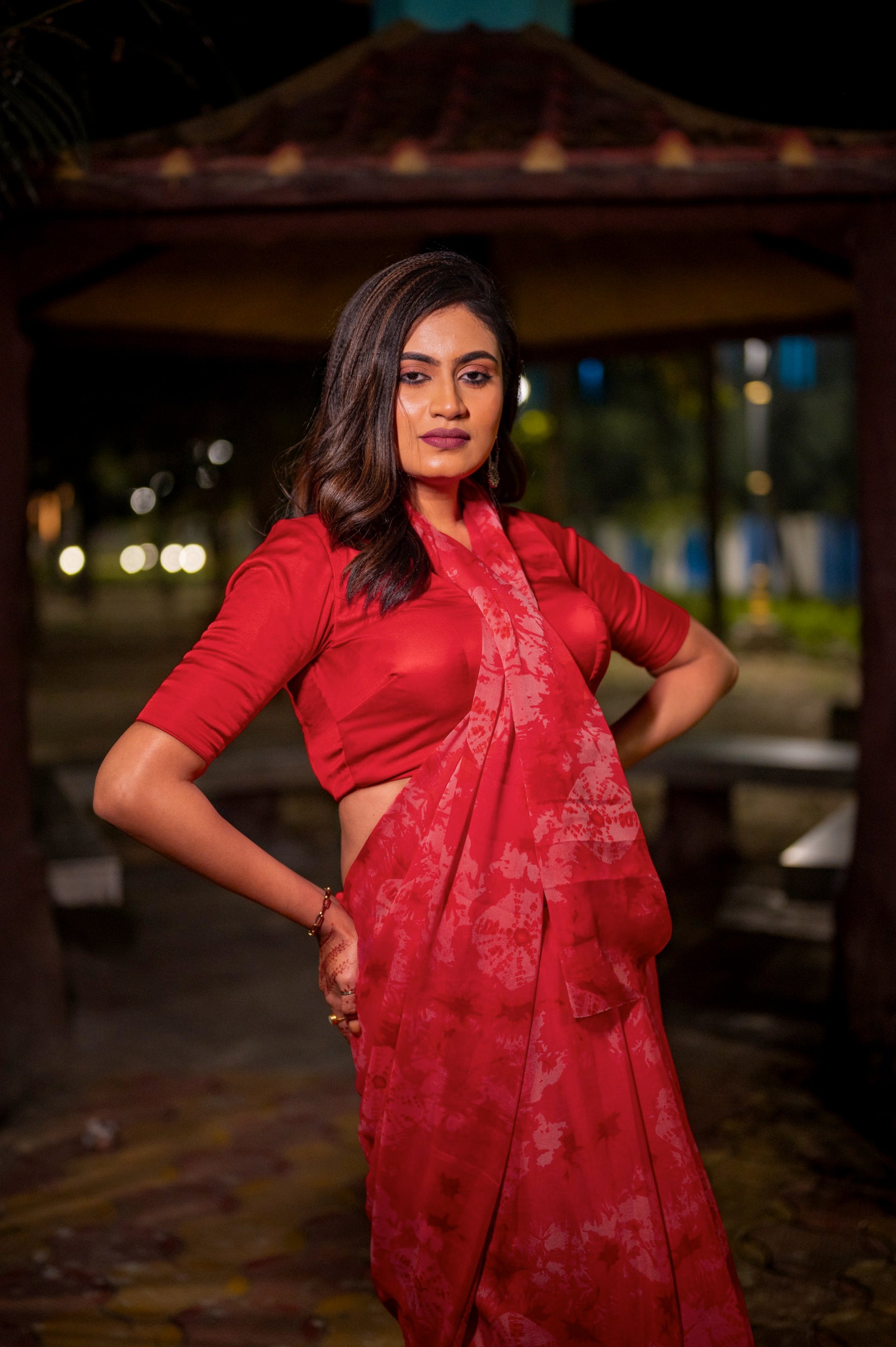 Woman wearing a rich red semi-silk blouse with front pleated detailing, styled with a matching red chiffon saree, posing confidently under soft outdoor lighting.