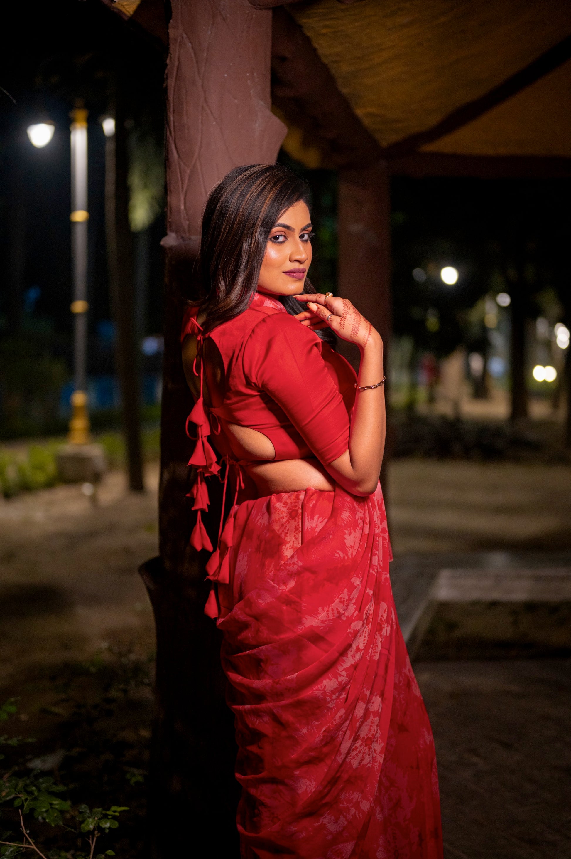 Side view of a red backless blouse with layered tie-up detailing, styled with a matching saree, captured in a softly lit outdoor space.