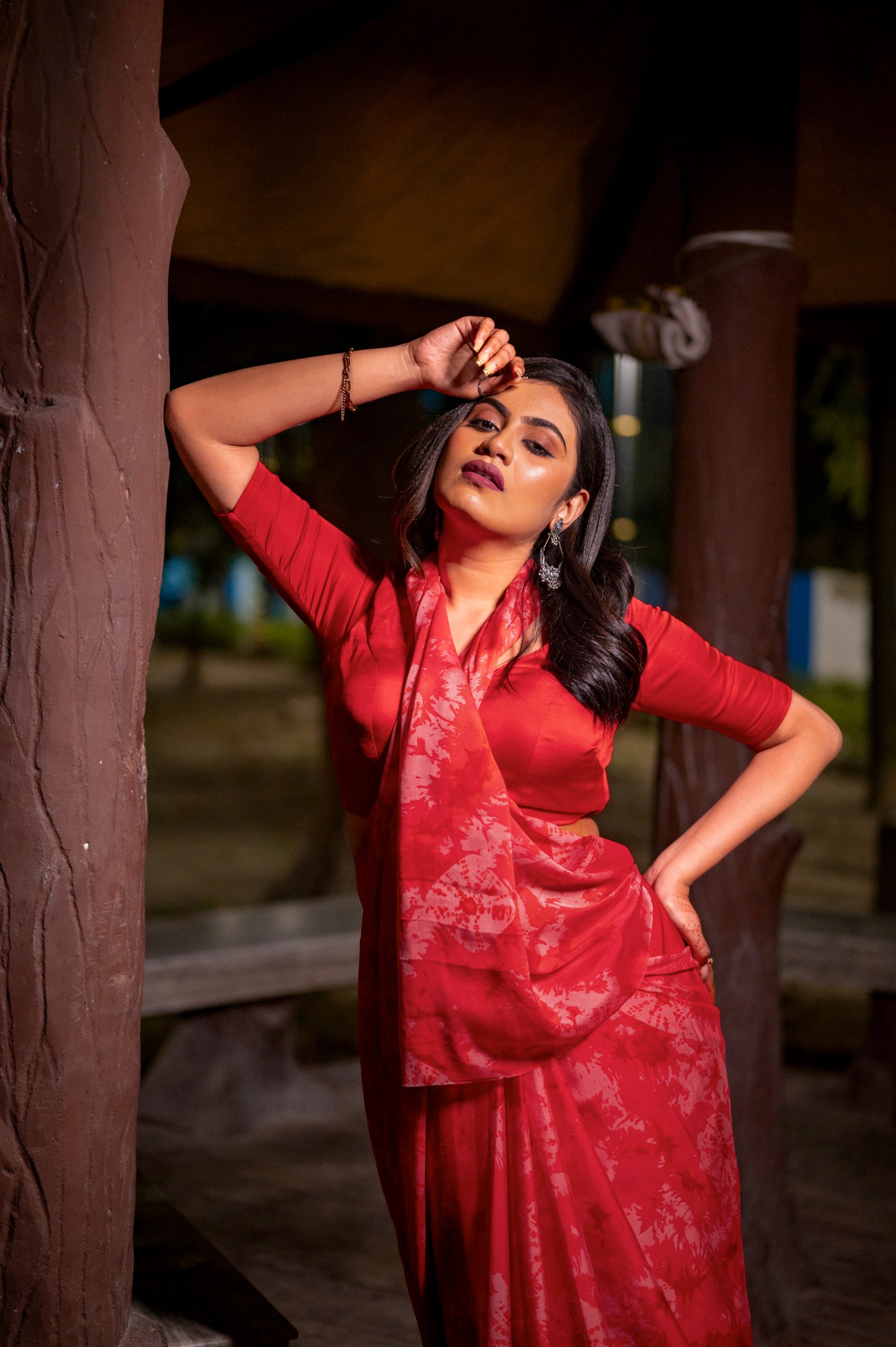Model in a striking red blouse and saree, leaning against a wooden pillar, exuding bold elegance in an outdoor nighttime setting.