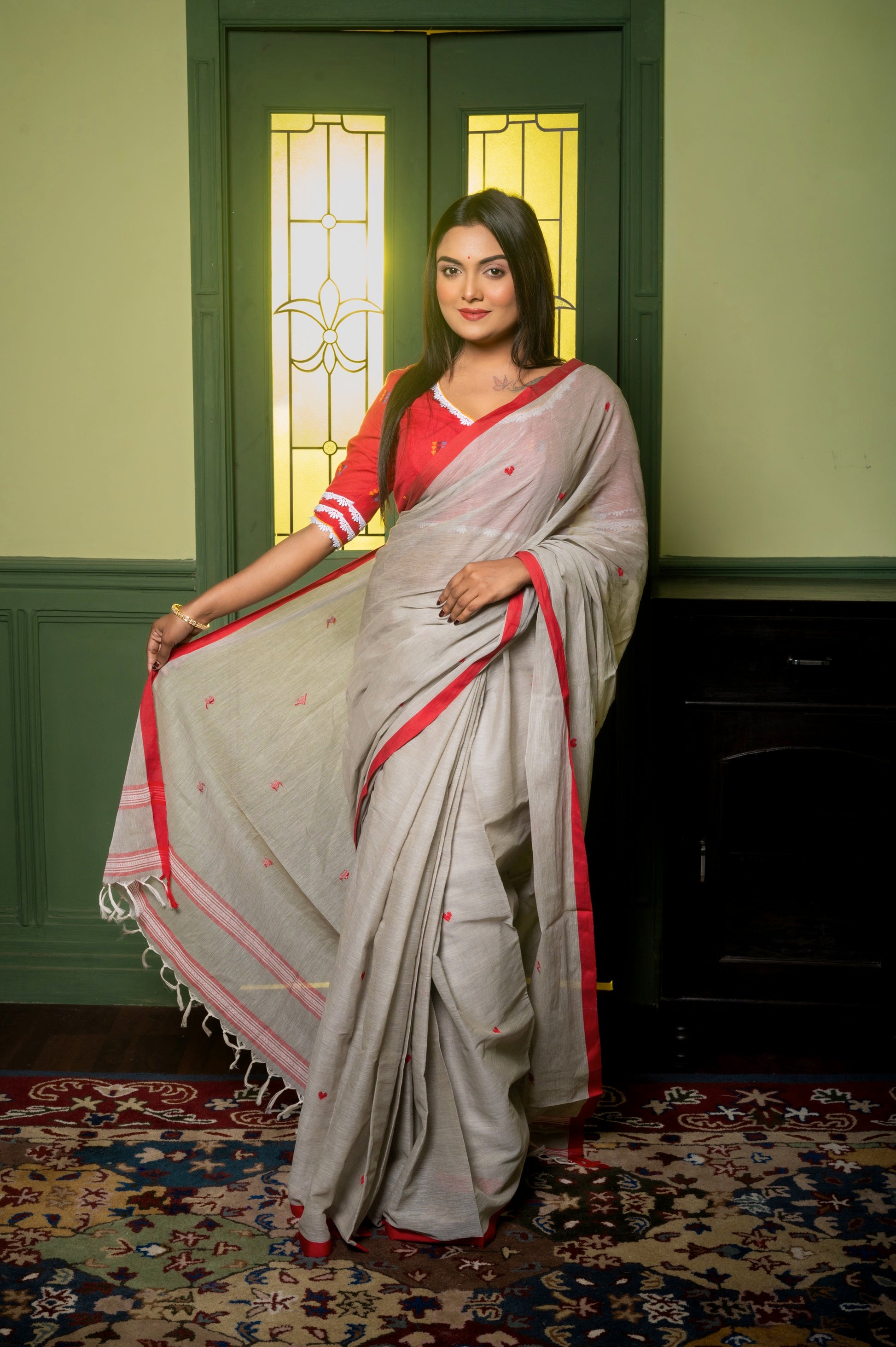 Full view of model in red cotton blouse and grey handloom saree with red border, standing confidently on a patterned rug in front of a traditional green doorway.