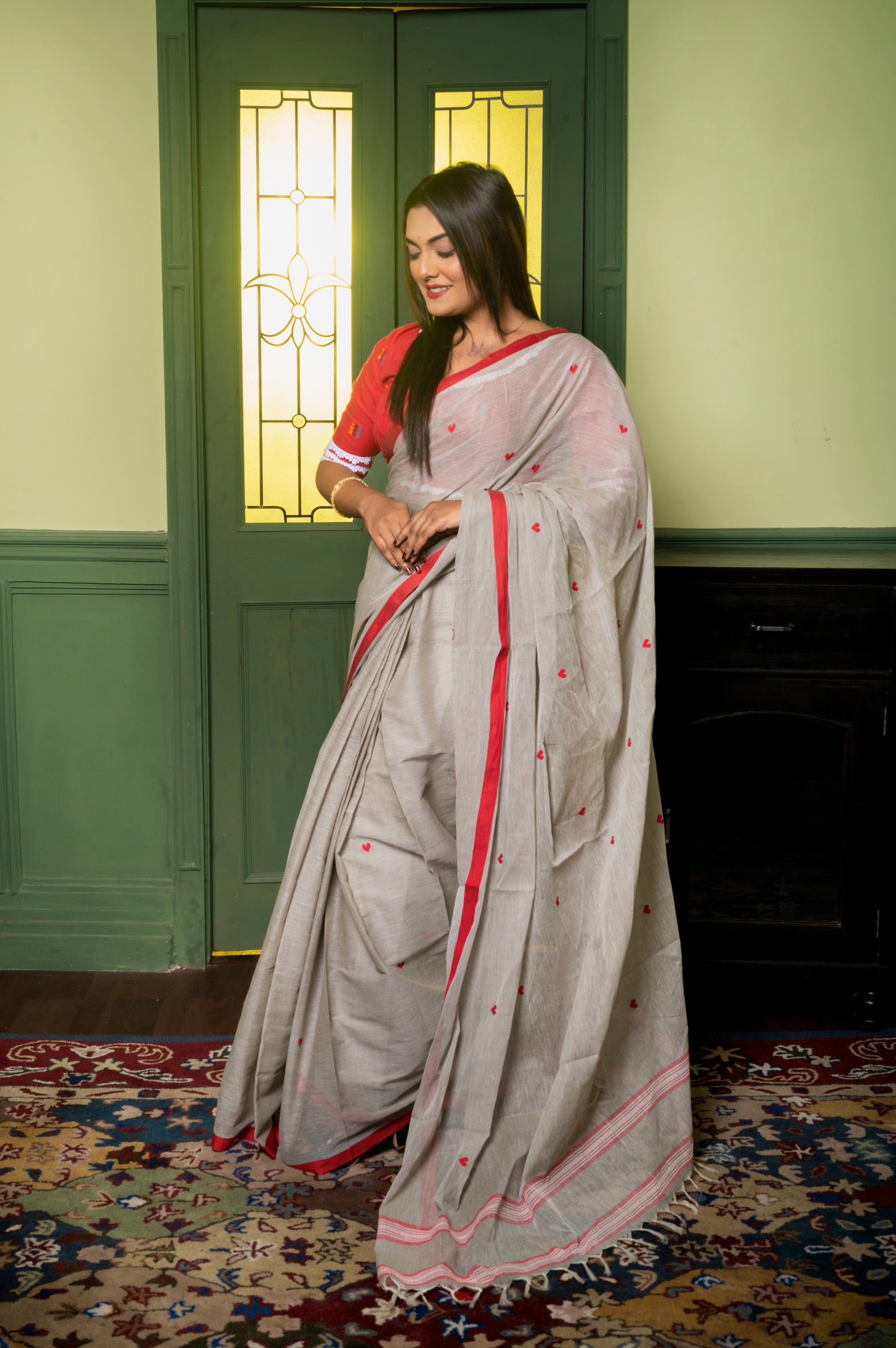 Model smiling in red blouse and grey saree with red piping, showcasing everyday ethnic elegance near a classic wooden door.