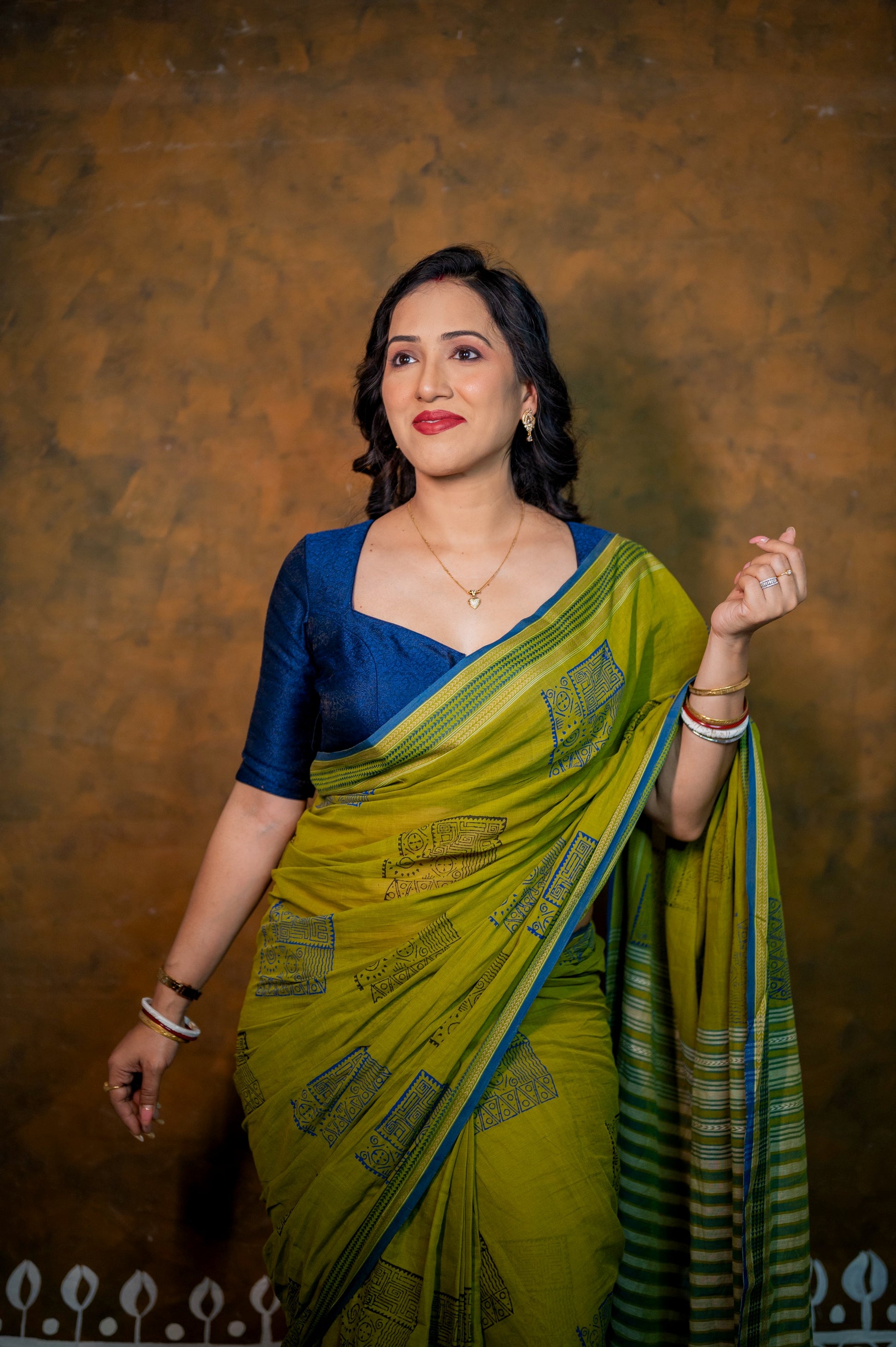 Woman wearing a bold blue jamebar blouse with square V neckline, paired with a green printed saree and traditional jewelry.