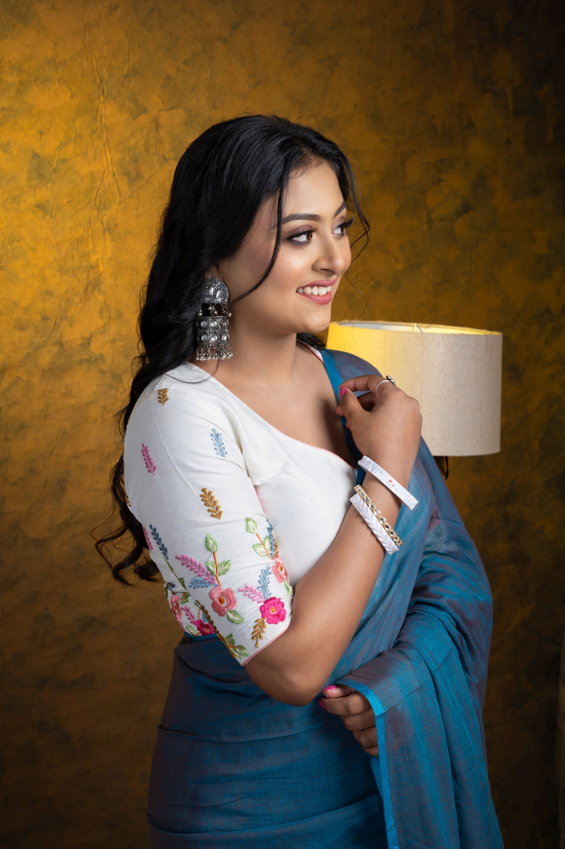 Woman wearing a white cotton-linen blouse with floral pastel embroidery on the sleeves, paired with a blue mulmul saree and statement jhumkas.