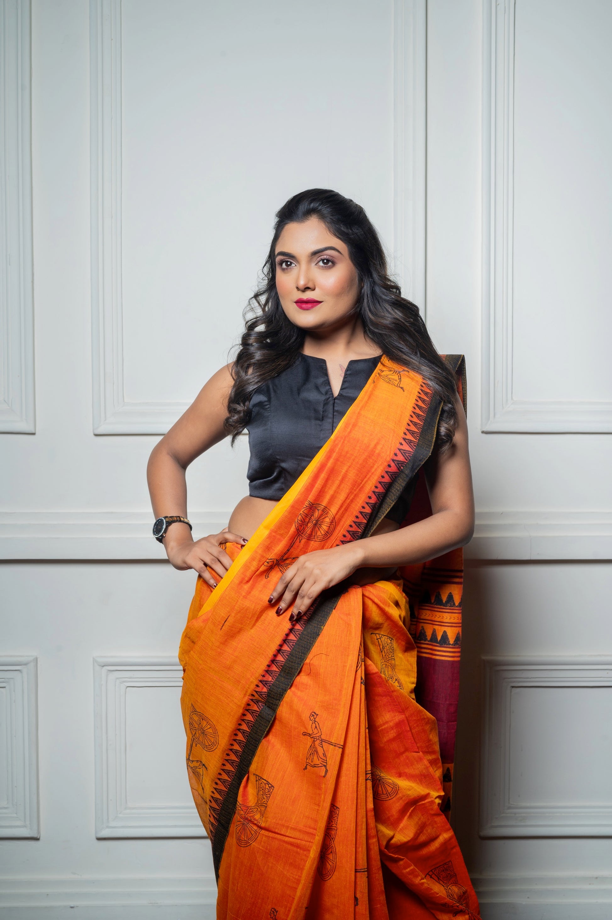 Woman wearing a sleeveless black semi-silk blouse with an O-neckline and an orange printed saree, posing confidently with one hand on hip.