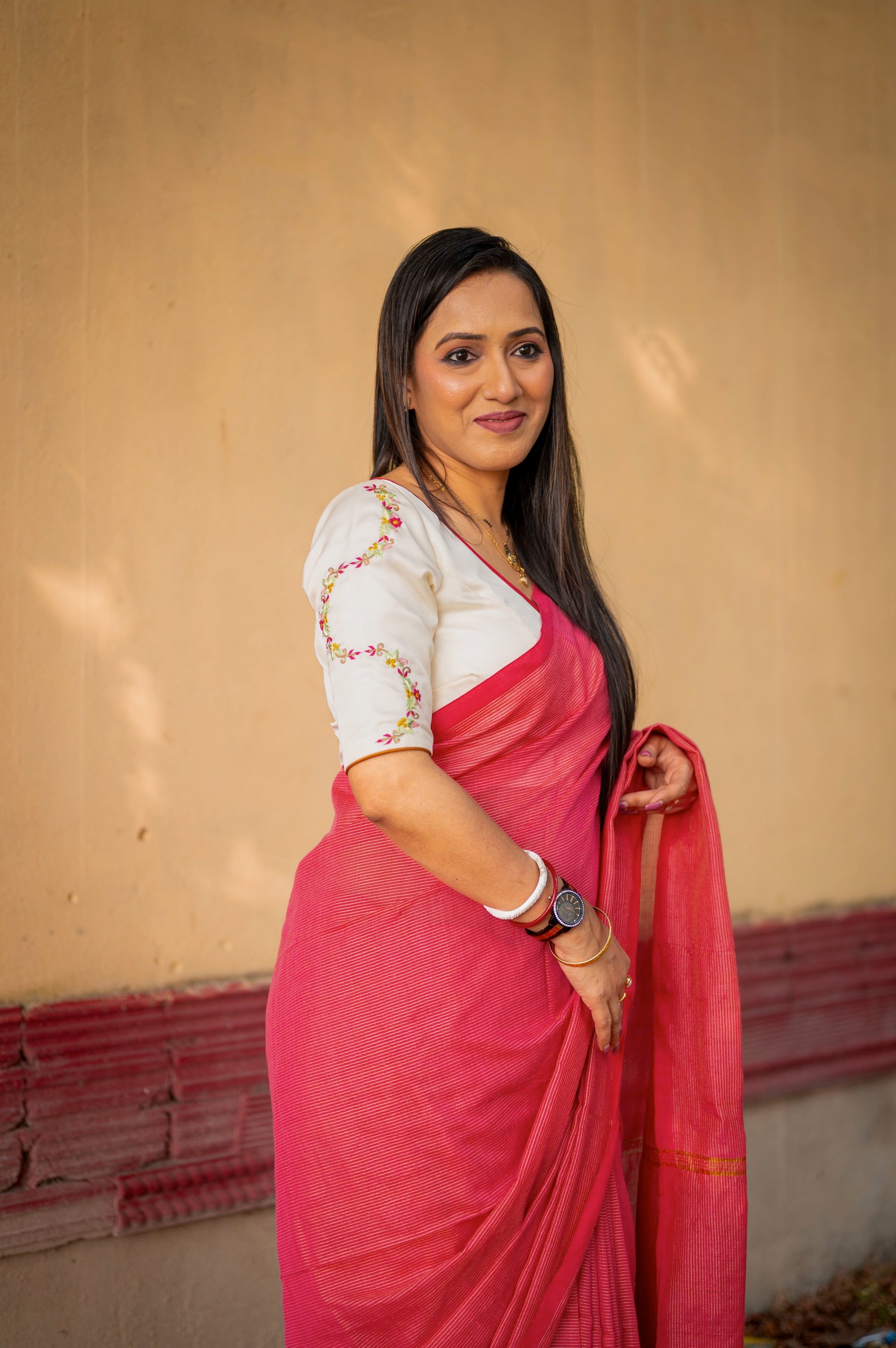 Model wearing an off-white semi-silk blouse with delicate pink and green embroidery on the sleeves, paired with a soft red handloom saree, posing against a warm beige wall.