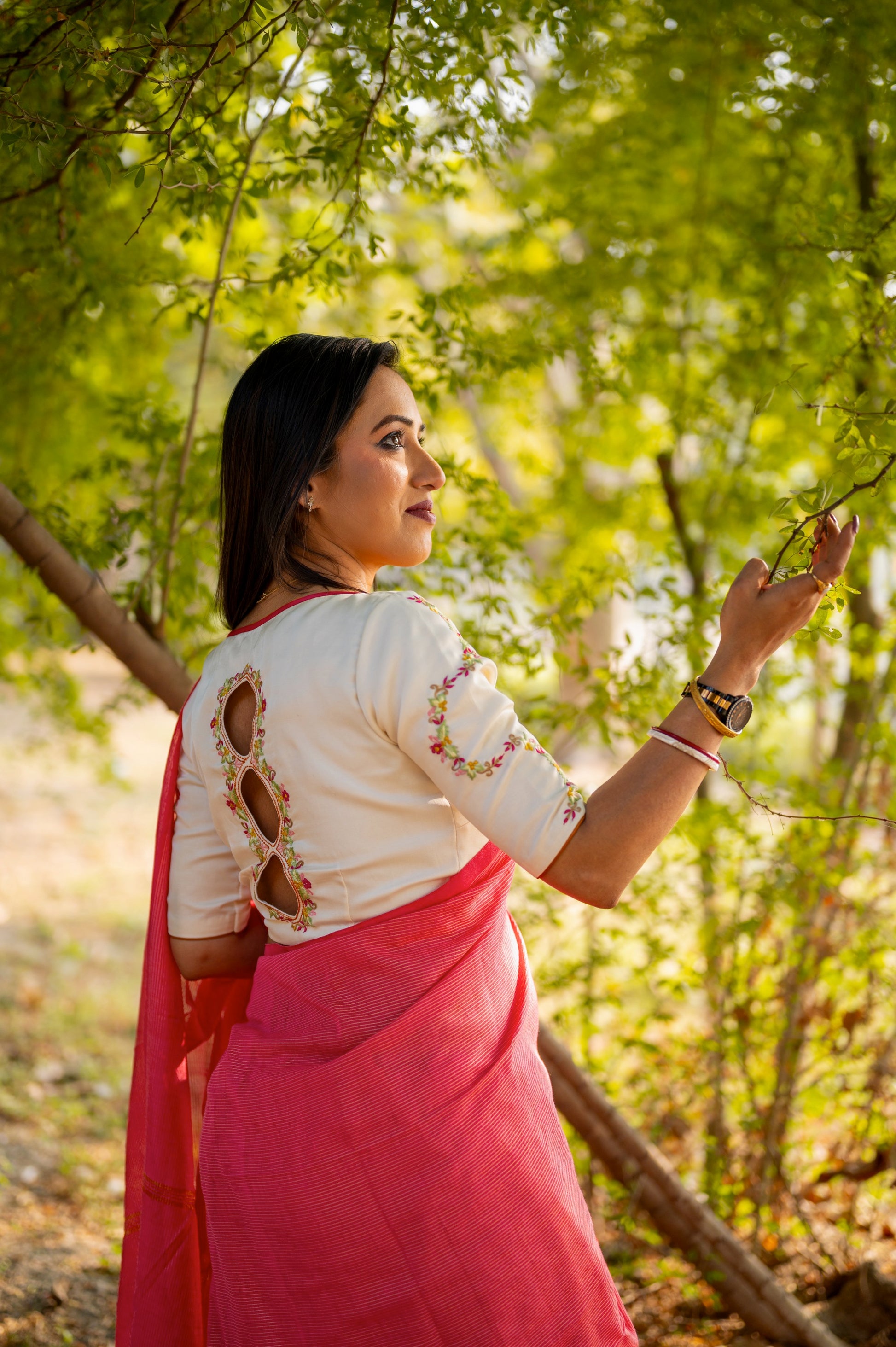 Model gently touching a tree branch while showcasing the back cutwork and embroidered sleeves of an off-white blouse, styled with a red handloom cotton saree.