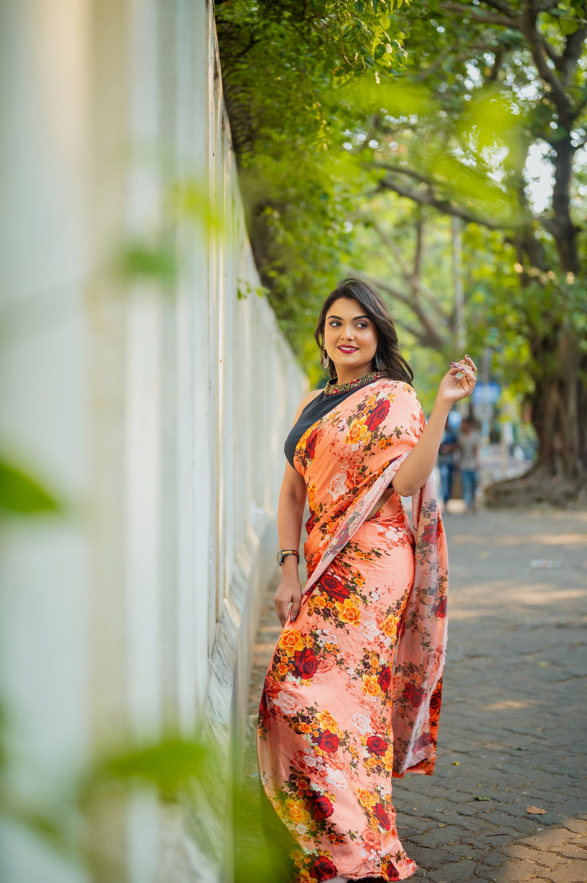 Woman wearing a black sleeveless halter-neck blouse with maroon and gold zardozi collar, paired with a floral peach saree on a sunlit sidewalk.