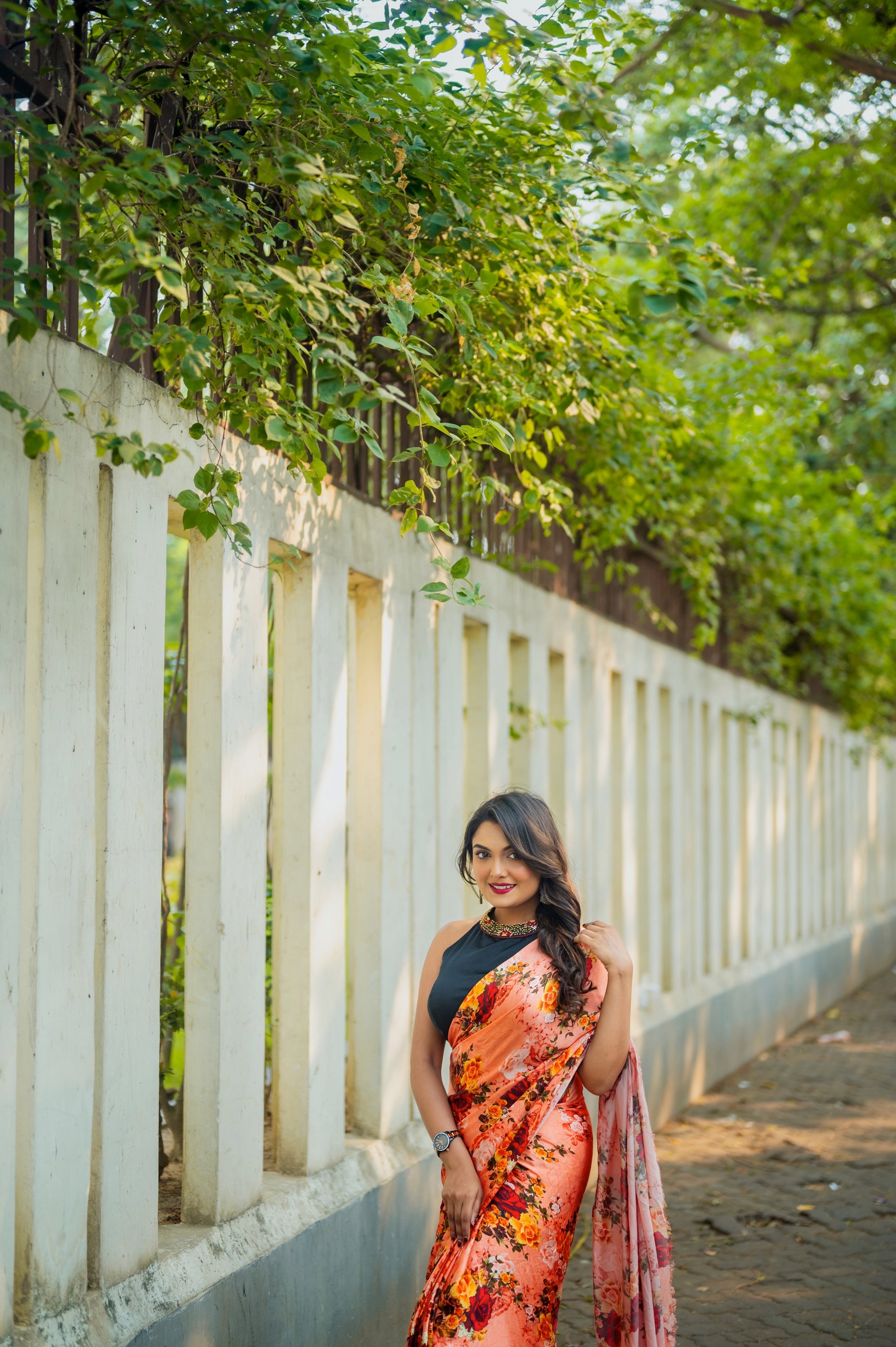 Full view of woman in black semi-silk halter blouse with maroon and gold collar, styled with peach floral saree under leafy arches.