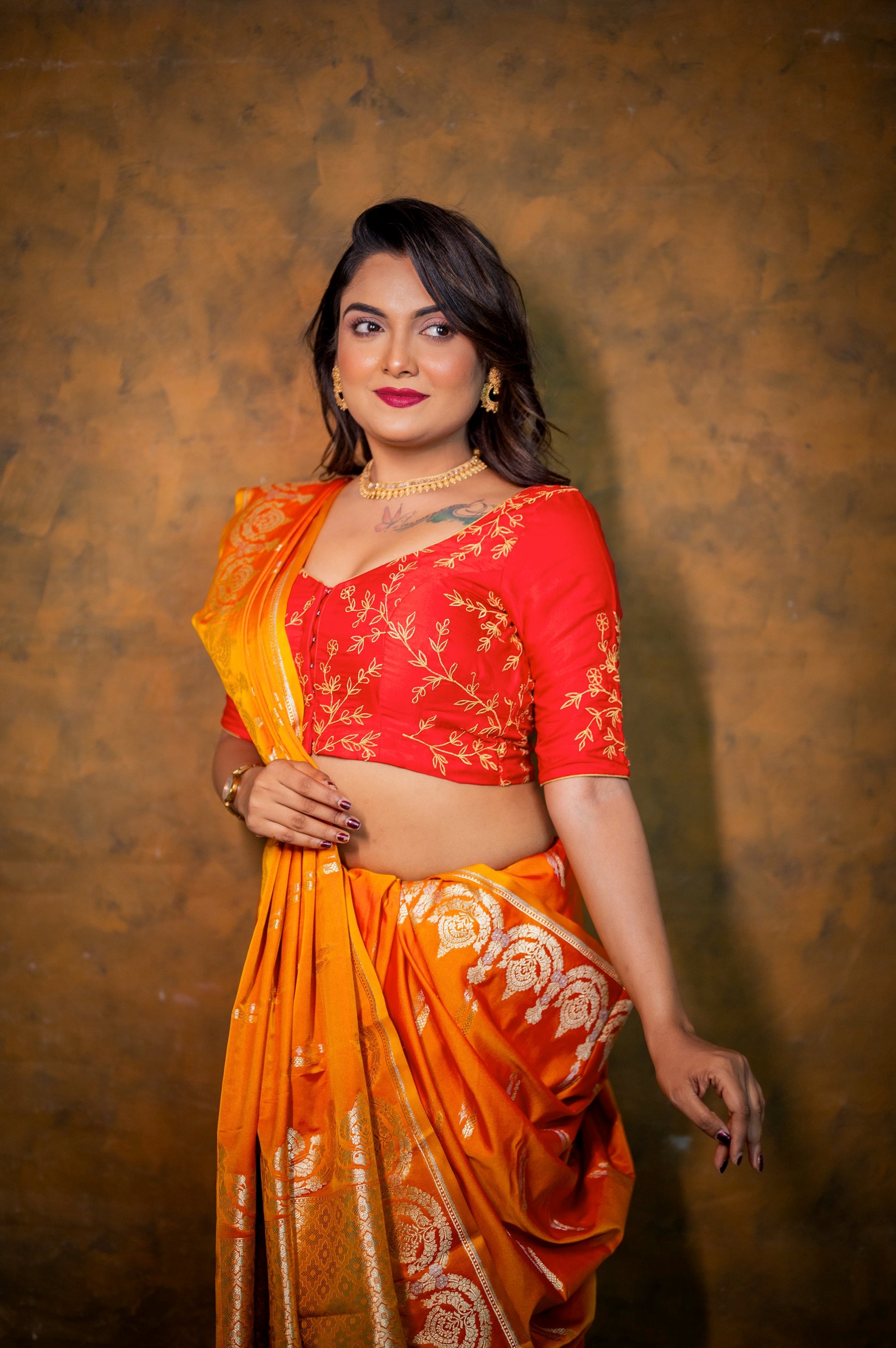 Woman wearing deep red semi-silk blouse with golden jari embroidery and wide V neckline, paired with vibrant orange saree.