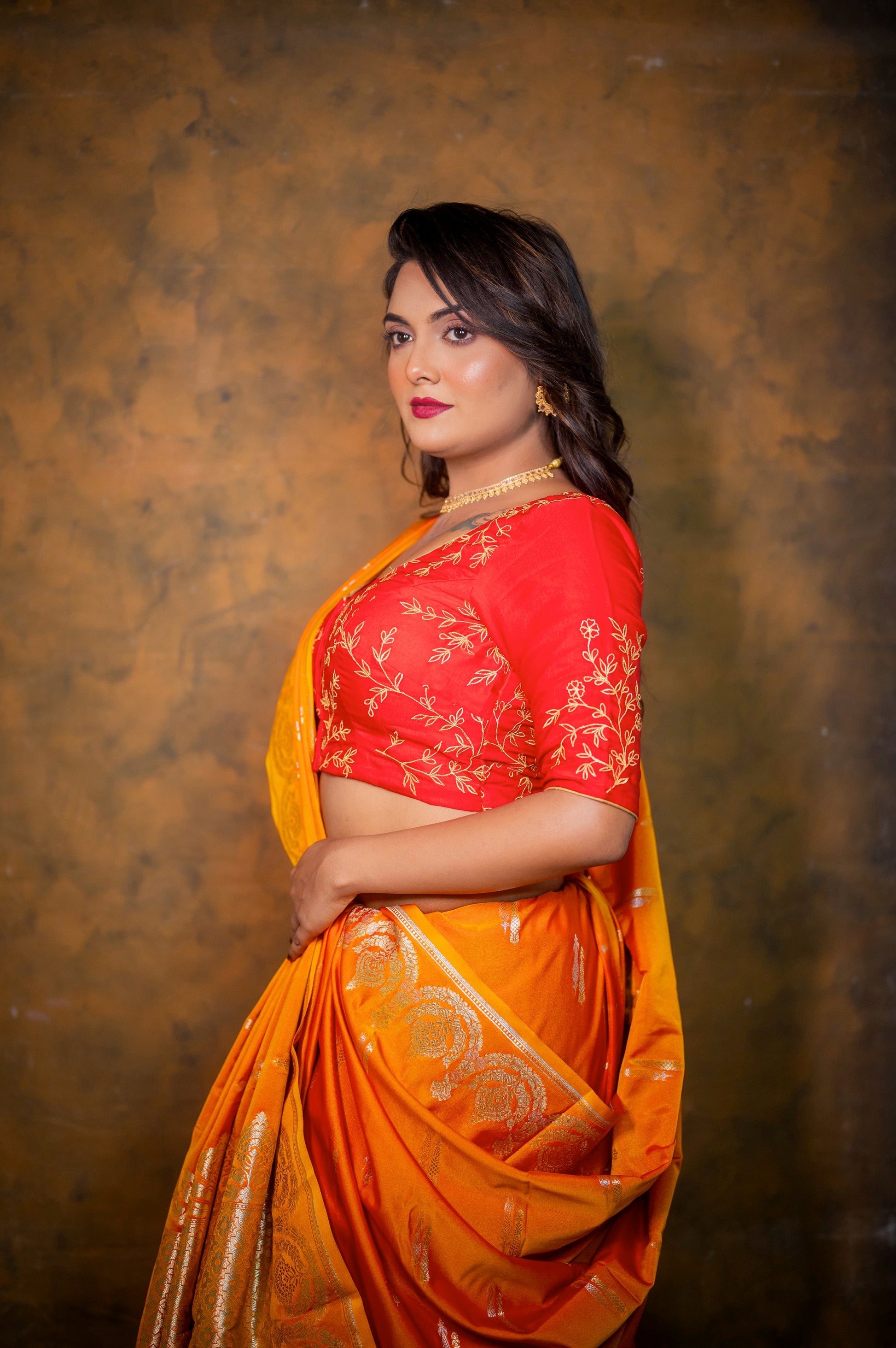 Side profile of woman in red embroidered blouse and orange saree highlighting the gold detailing and tailored fit.
