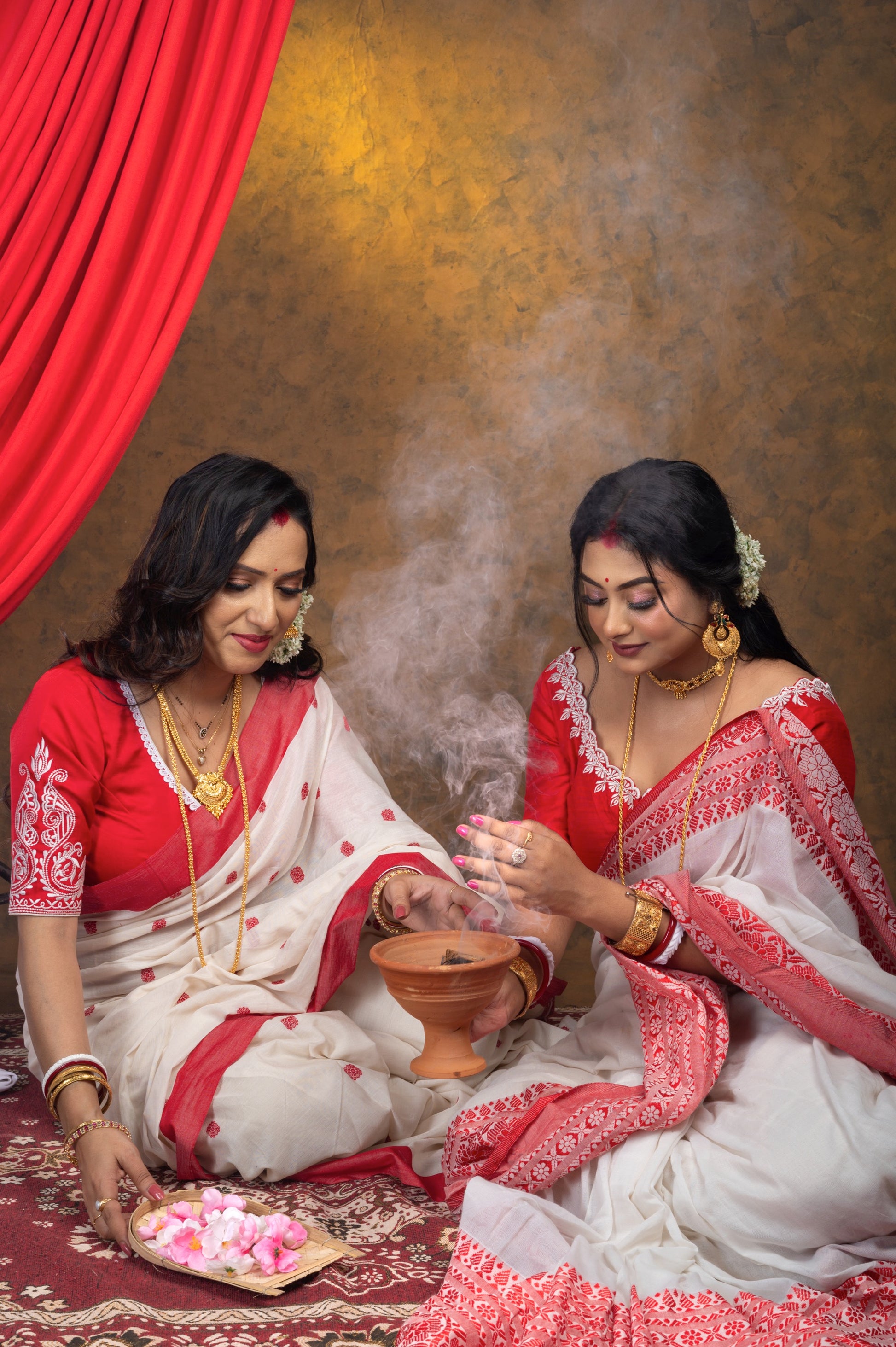Close-up of red blouse with off-white embroidery on neckline and sleeves, styled with red and white jamdani saree, two women performing traditional Bengali puja with incense smoke