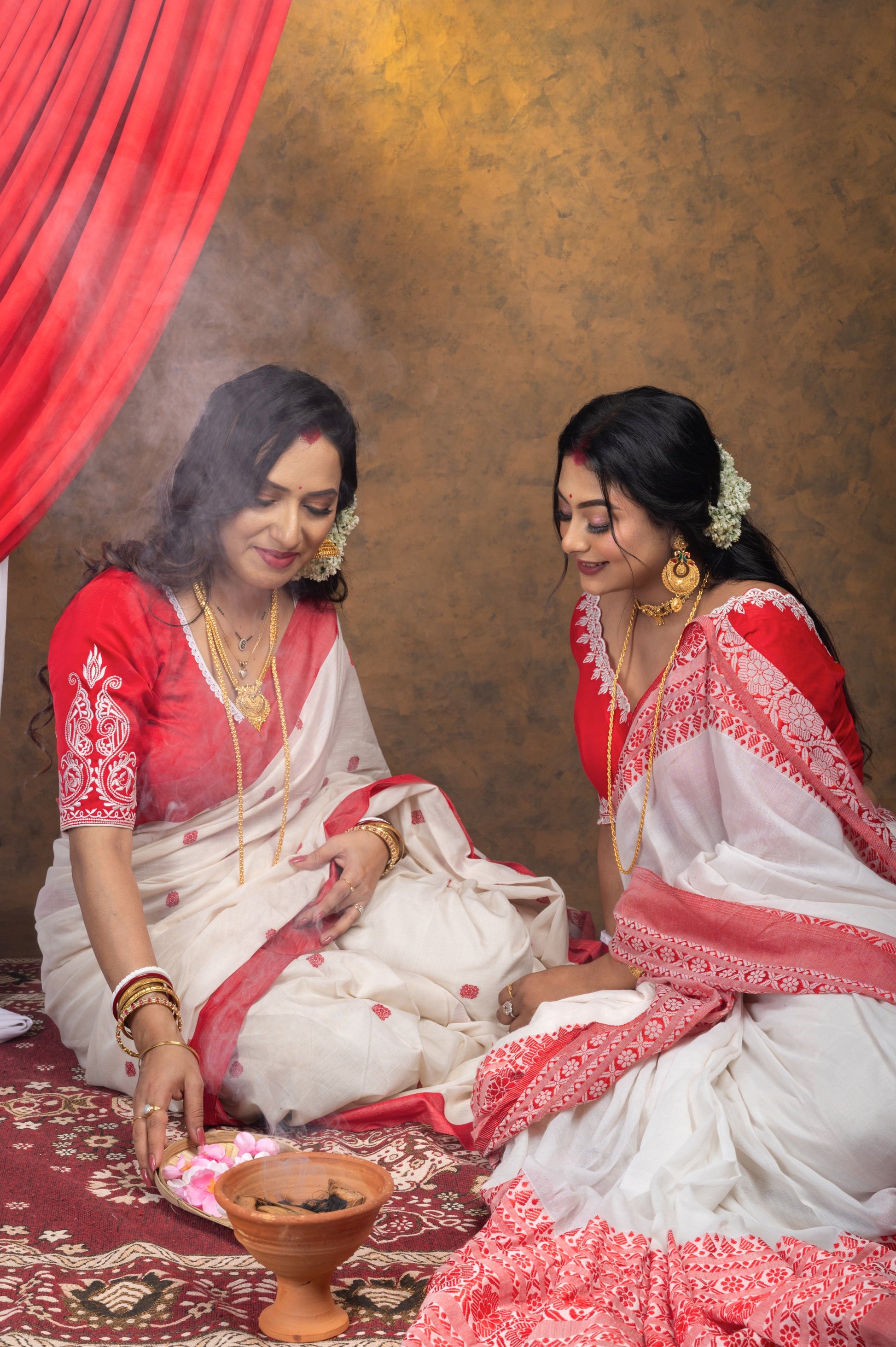 Two women dressed in traditional Bengali red-bordered sarees, both wearing red semi-silk blouses with white embroidery, engaged in a puja ceremony surrounded by incense and flowers