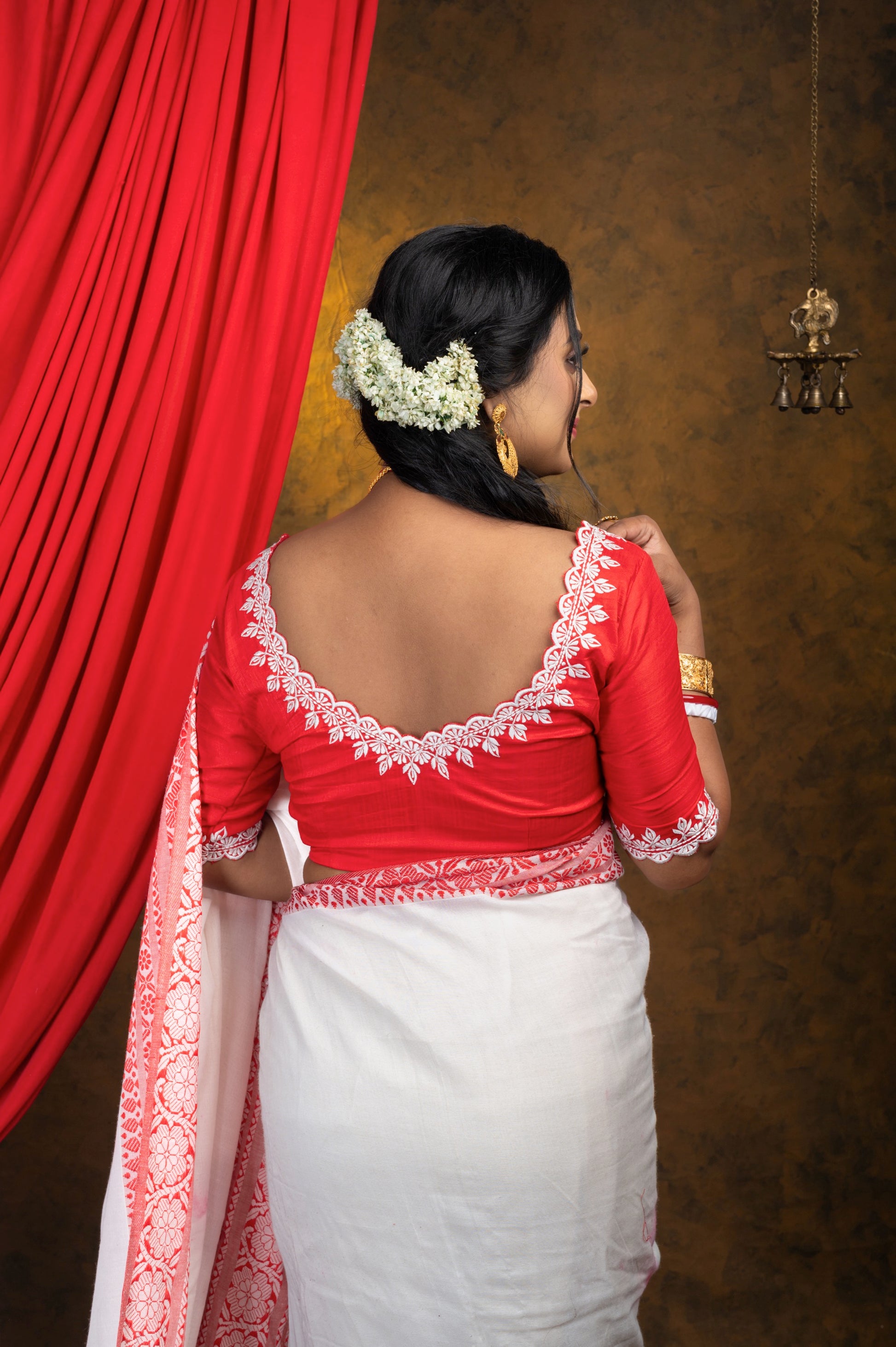 Back view of red semi-silk blouse featuring wide V neckline and off-white leaf-pattern embroidery, paired with a traditional white saree with red borders and gajra-adorned hair