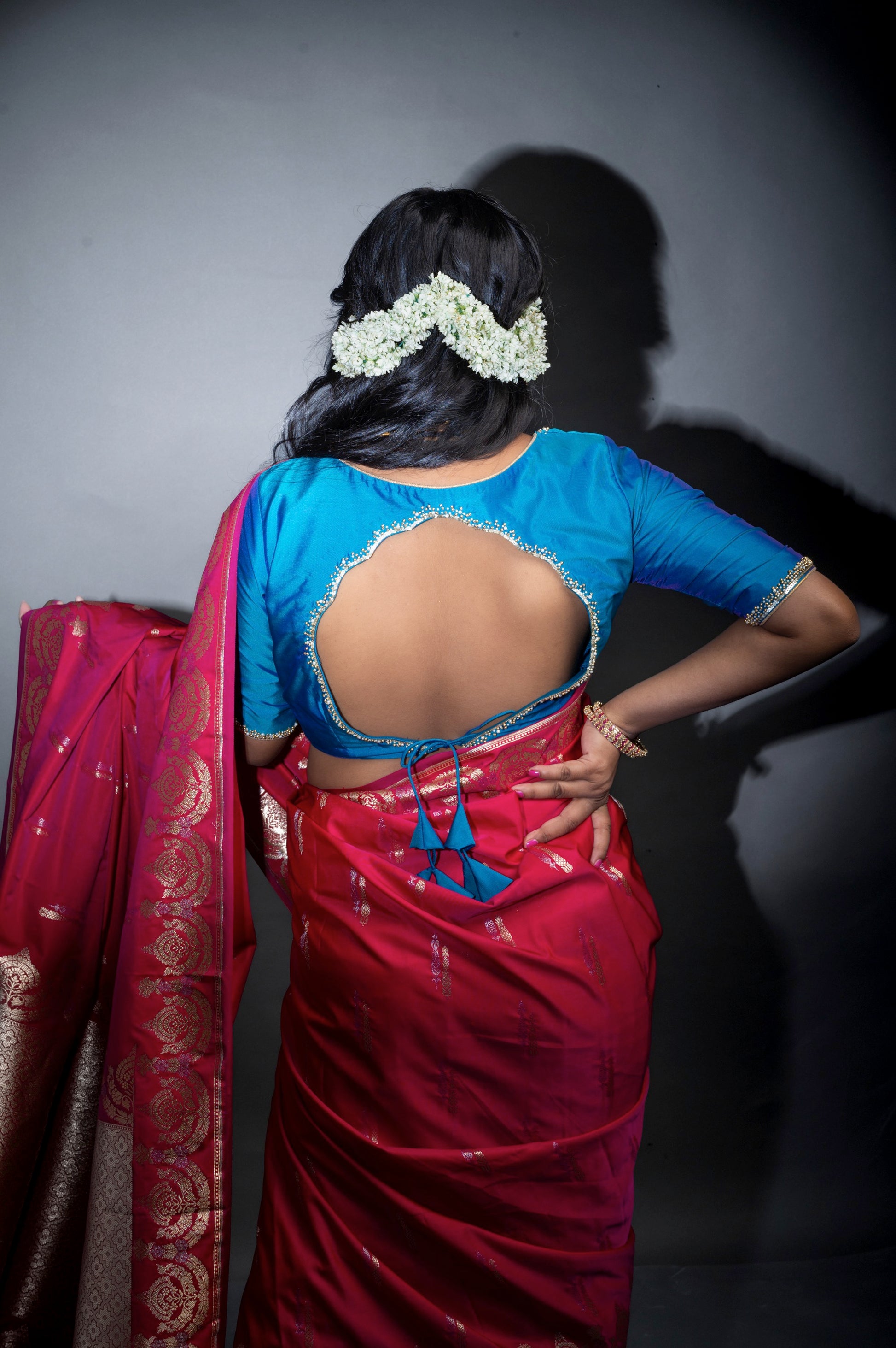 Back view of a designer teal blue blouse featuring a deep U-shaped cut and embellished scallop edges, tied with dual tassels, styled with a rich red silk saree.