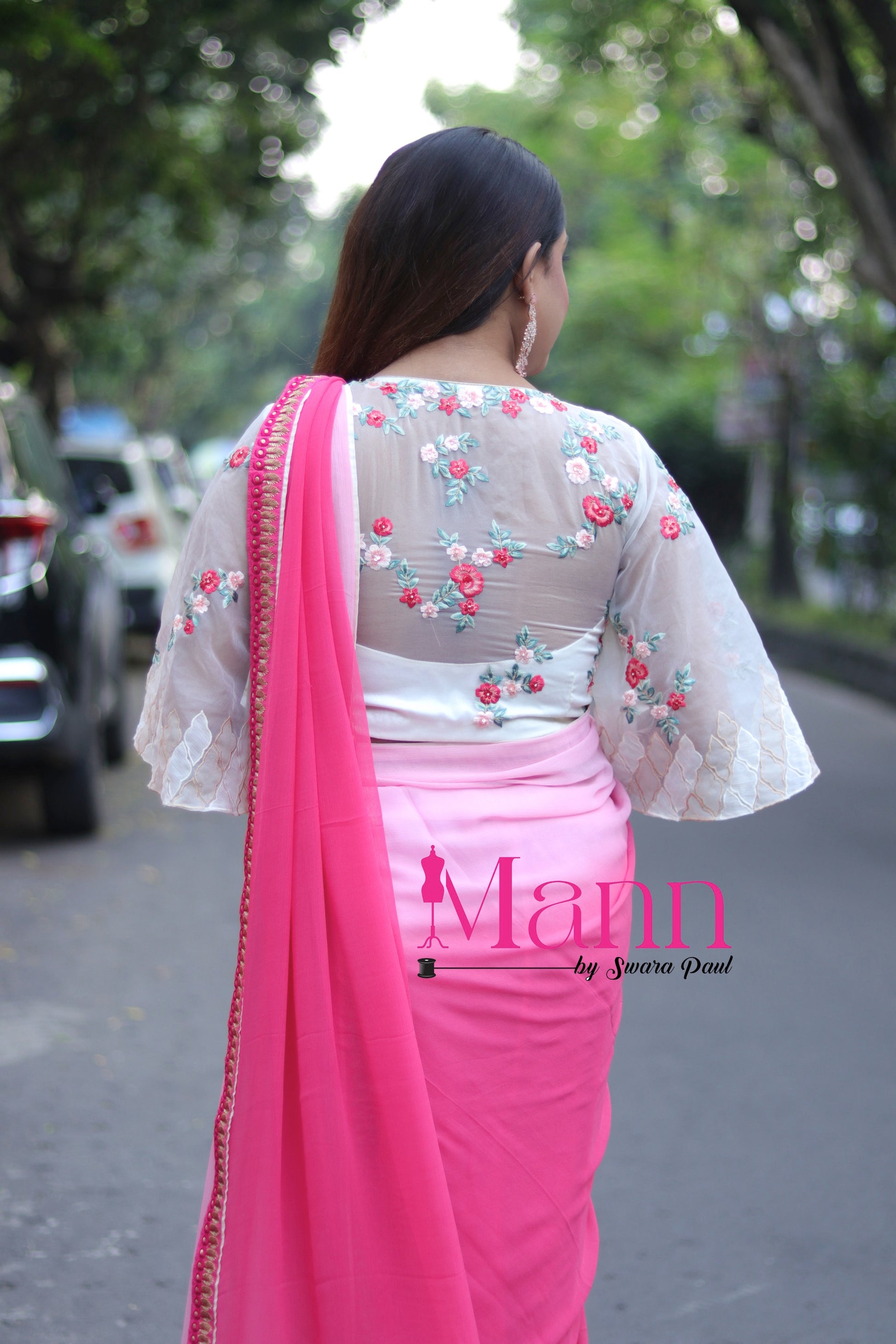 Back view of white semi-silk blouse with sheer panel fully embroidered with pink and green floral threadwork, paired with a pink saree featuring decorative border, model walking down the road