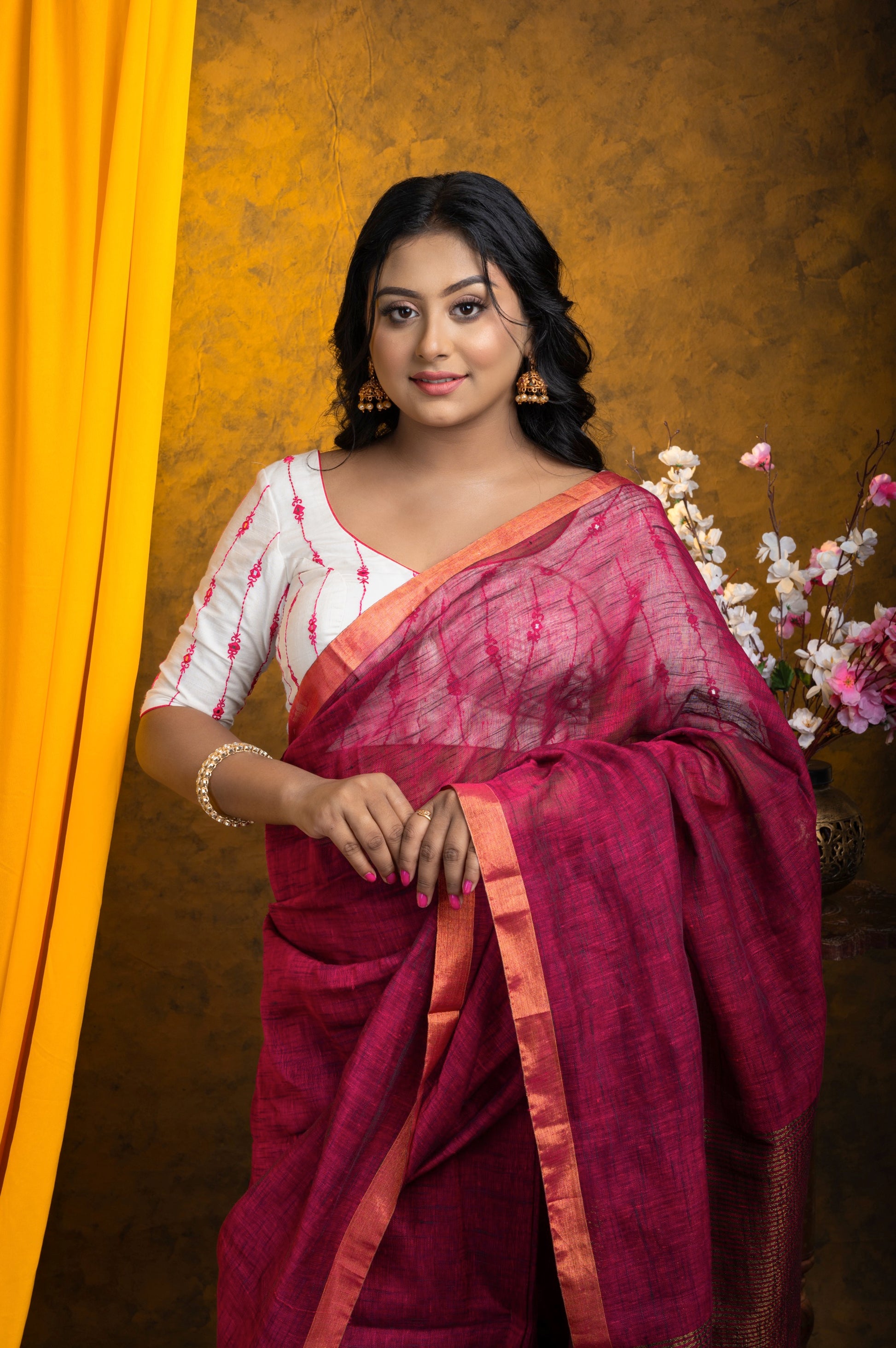 A woman wearing a stylish white designer blouse with colorful embroidery, elbow-length glass sleeves, and a deep V-neckline, posing confidently against a beige background