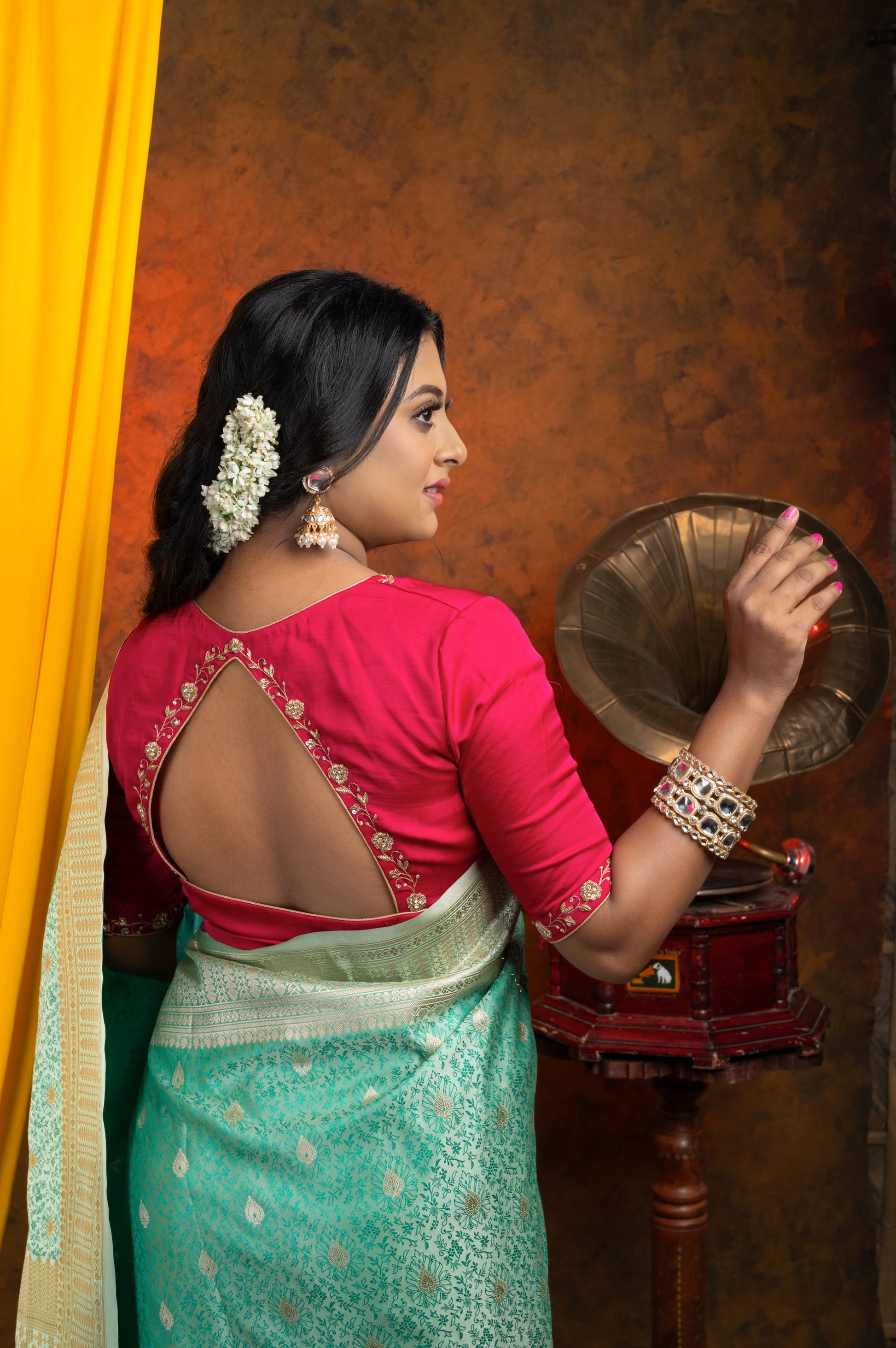 Back view of rani pink semi-silk blouse featuring a diamond cut-out with gold zardozi embroidery and white stone detailing, styled with a mint green Banarasi saree and traditional accessories