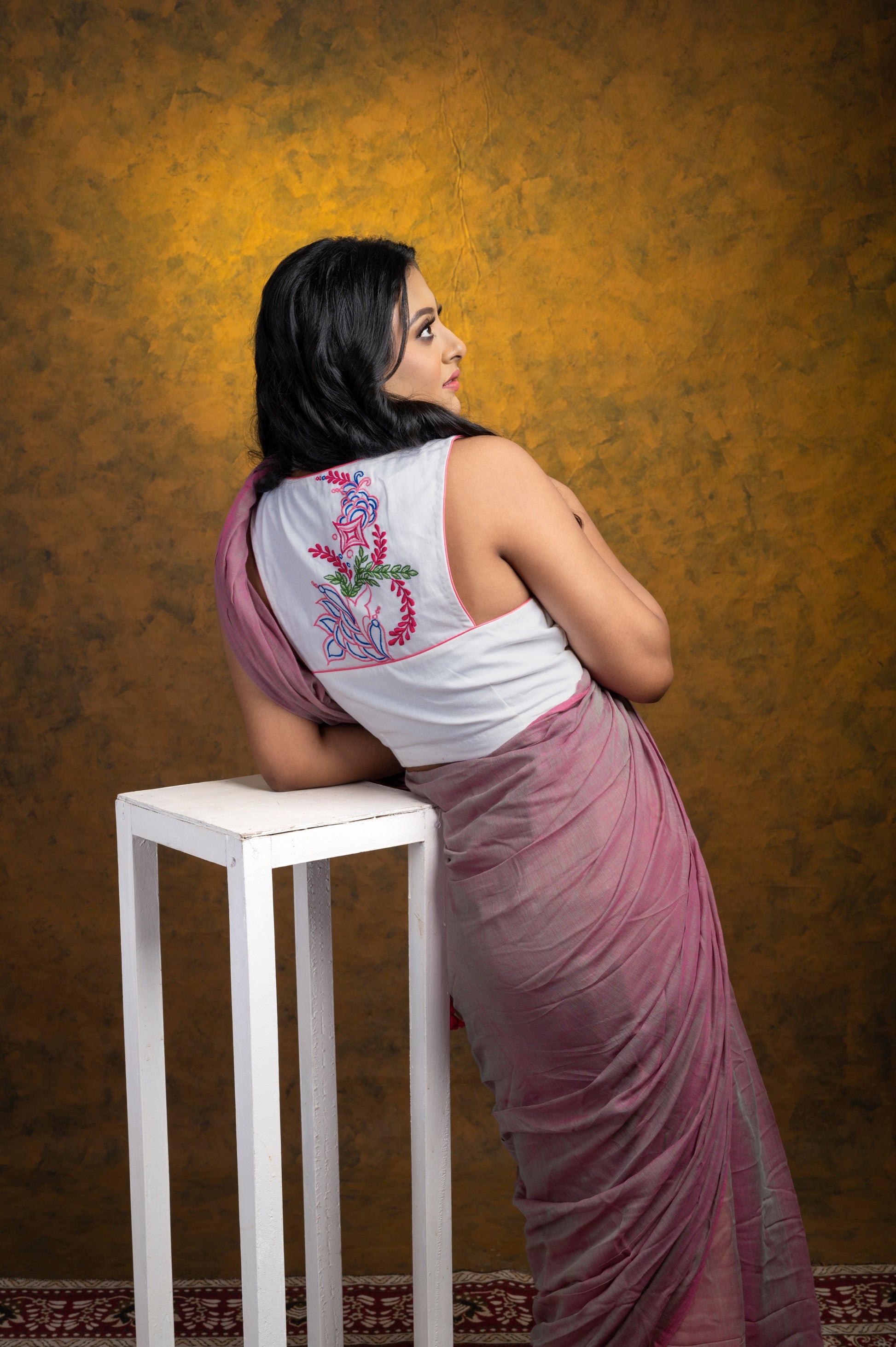 Back view of white sleeveless cotton-linen blouse with pastel pink piping and multicolor floral embroidery, paired with a mauve saree, model leaning against a white wooden stand