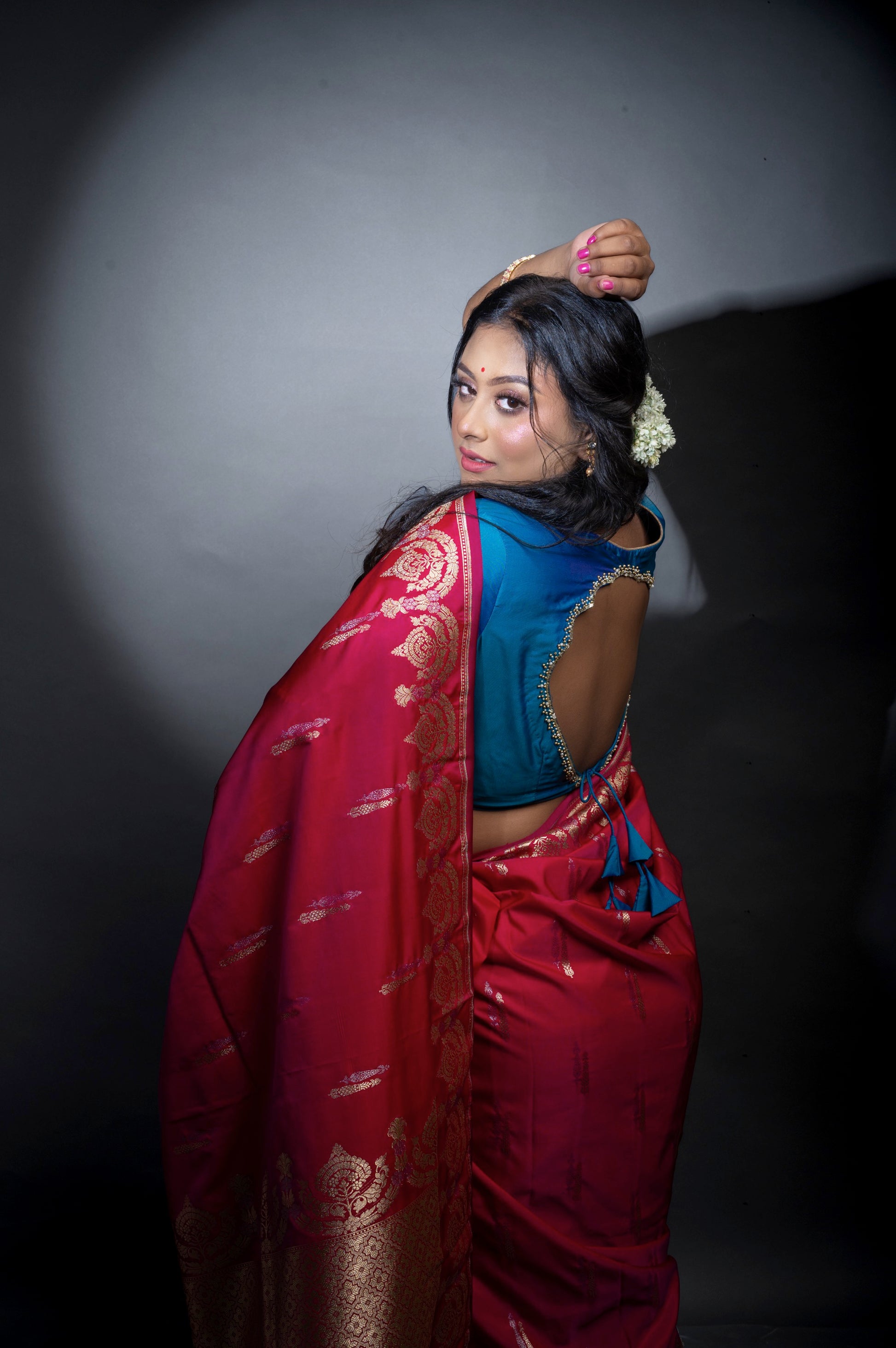  Close-up side view of a woman in a deep teal blouse with shimmering sleeve borders and plunging neckline, paired with a contrasting red saree for an elegant festive look.