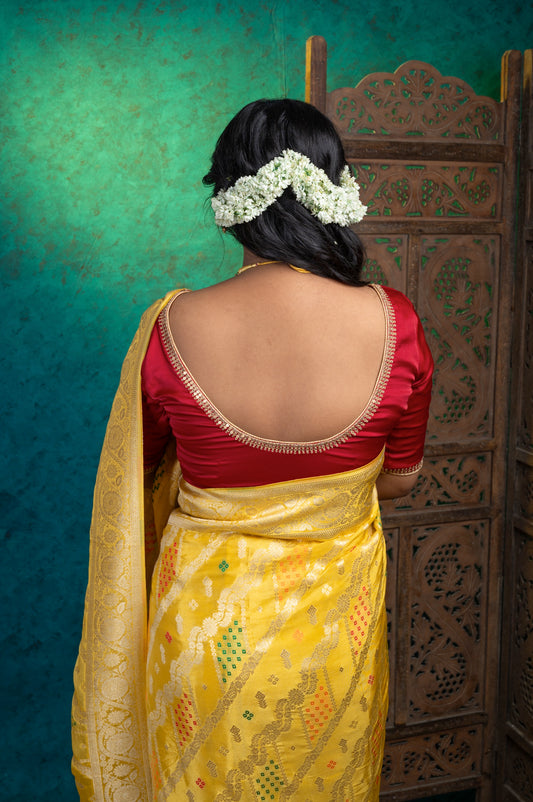 Woman in a yellow Banarasi saree showcasing the deep-cut back of a rich maroon semi-silk blouse with delicate hand-done zardozi embroidery, styled with gajra-adorned hair.